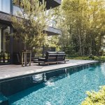 ThumbnailFotografie van een moderne villa met een strak ontwerp, blauw zwembad reflecterend licht, grijs houten terras met loungeset, omgeven door groen en zonnestralen.