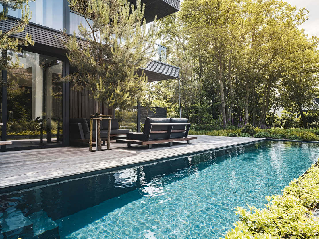 Fotografie van een moderne villa met een strak ontwerp, blauw zwembad reflecterend licht, grijs houten terras met loungeset, omgeven door groen en zonnestralen.