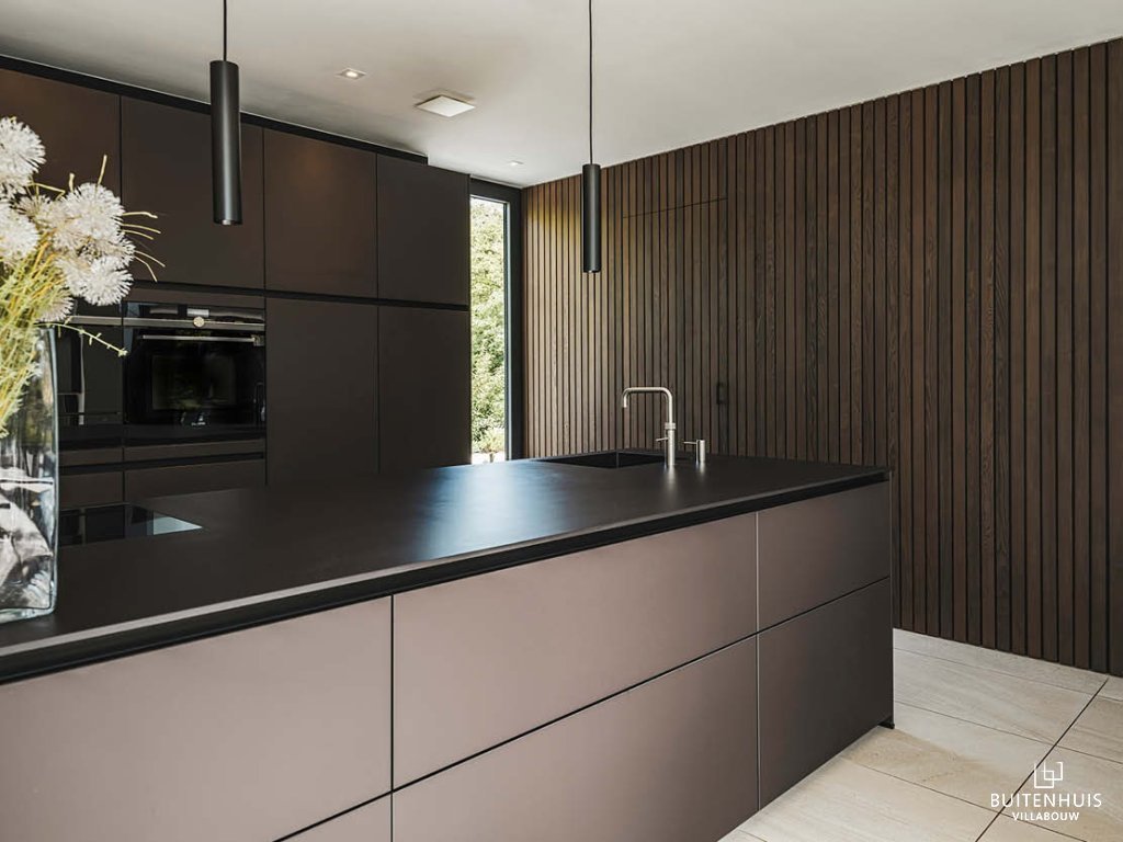 ThumbnailInterieurfoto van een moderne keuken met eiland, donkere kastfronten, zwarte hanglampen en houten wandbekleding met verticale lijnen.