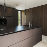 ThumbnailInterieurfoto van een moderne keuken met eiland, donkere kastfronten, zwarte hanglampen en houten wandbekleding met verticale lijnen.