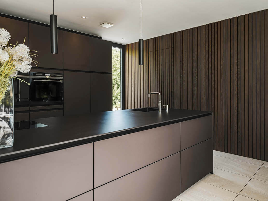 Interieurfoto van een moderne keuken met eiland, donkere kastfronten, zwarte hanglampen en houten wandbekleding met verticale lijnen.