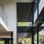 ThumbnailInterieurfoto van een modern huis met glazen balustrade, grote ramen met rolgordijnen en lichtinval, met deel van een metalen booglamp.