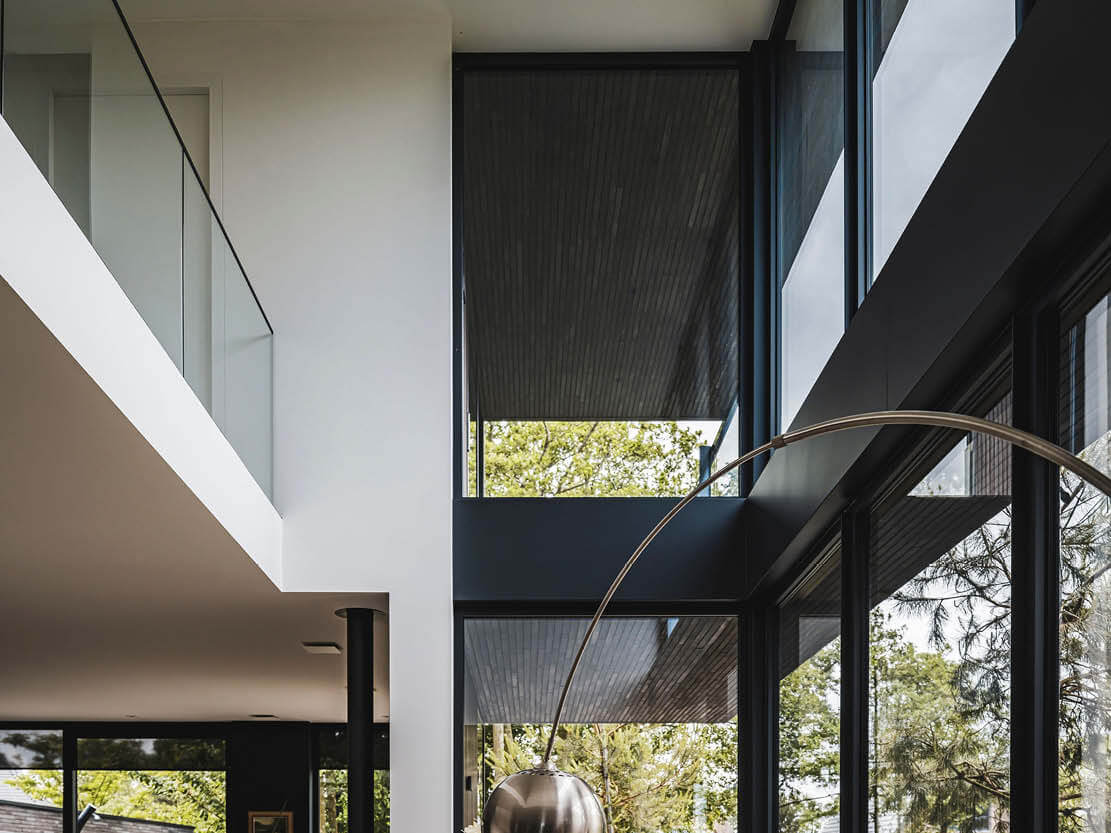 Interieurfoto van een modern huis met glazen balustrade, grote ramen met rolgordijnen en lichtinval, met deel van een metalen booglamp.