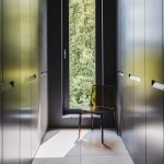 ThumbnailFoto van een smalle ruimte met donkere kasten die goud lijken te kleuren door lichtinval, stoel bij raam met uitzicht op groen, lichte tegelvloer.