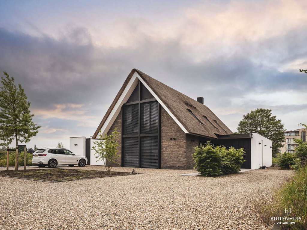 ThumbnailVrijstaand huis met rieten dak, grote ramen, een witte SUV auto op de oprit, grindpad en bomen.