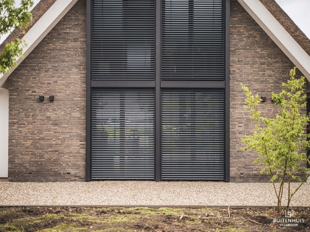 ThumbnailFoto van een bakstenen huis met grote, grijze ramen/deuren en een traditioneel rieten dak, met grindpad en planten in de voortuin.