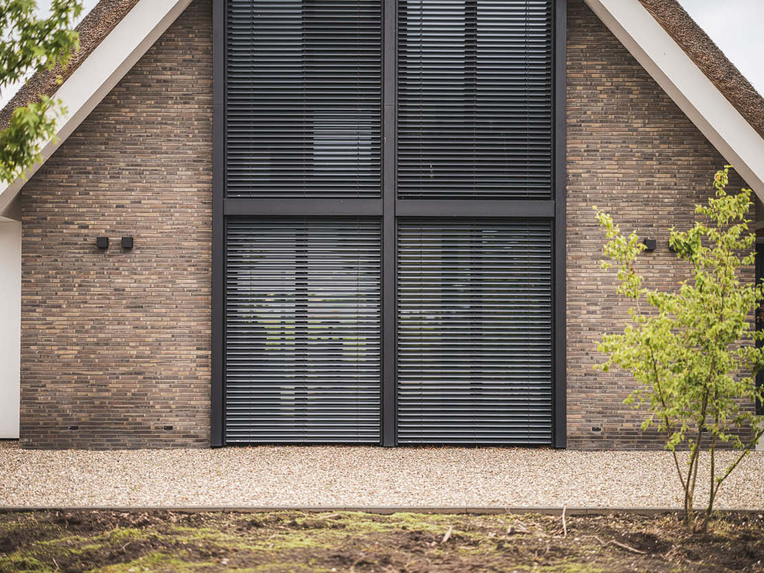 Foto van een bakstenen huis met grote, grijze ramen/deuren en een traditioneel rieten dak, met grindpad en planten in de voortuin.
