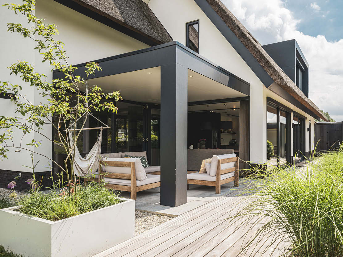 Buitenfoto van een rietgedekt huis met een modern terras, houten zitmeubels en schaduwrijke veranda.