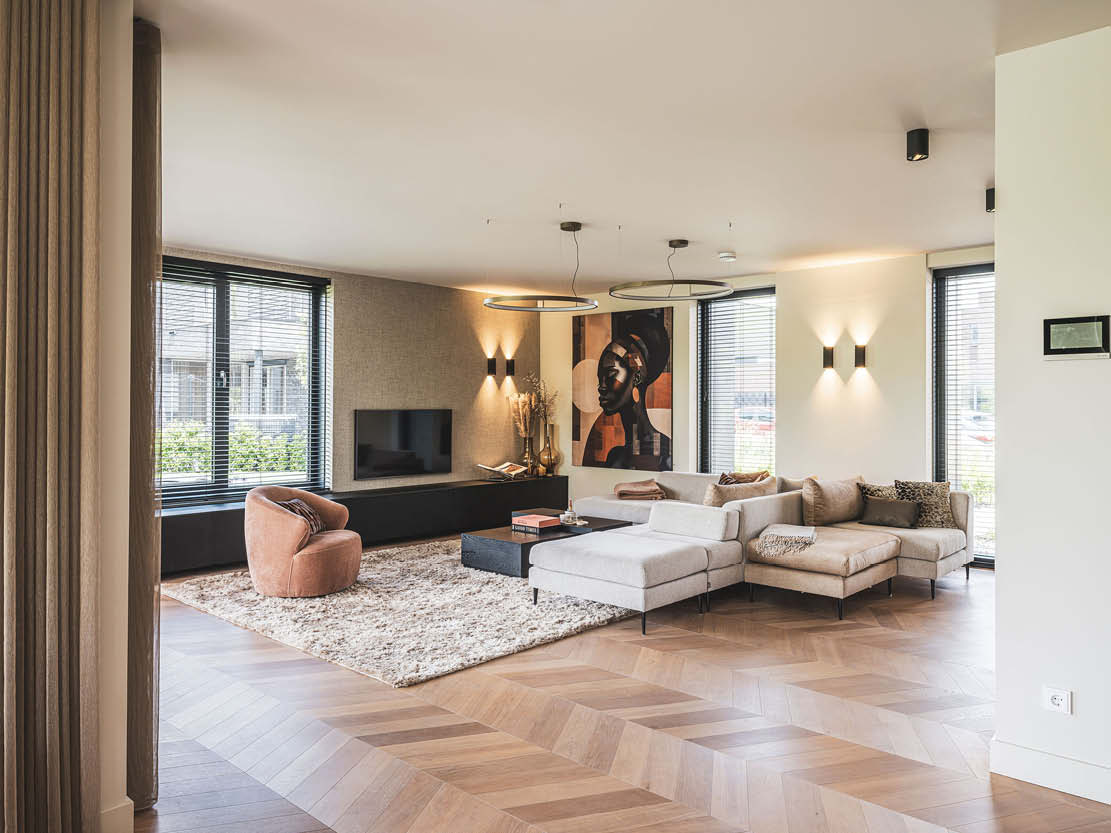 Interieurfoto van een moderne woonkamer met een opvallend kunstwerk aan de muur, een hoekbank, fauteuil en een houten parketvloer.