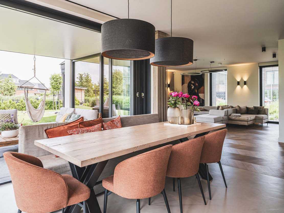Interieurfoto van een woonkamer met een houten eettafel met oranje stoelen, grijze hanglampen, bank met kussens en grote schuifdeuren.