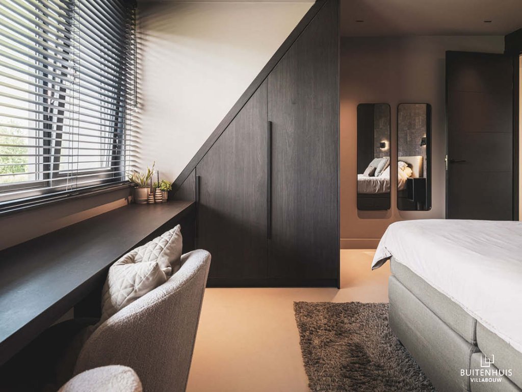 ThumbnailInterieurfoto van een slaapkamer met donkere op maat gemaakte kast onder hellend plafond, raam met jaloezieën en grijze boxspring.