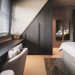ThumbnailInterieurfoto van een slaapkamer met donkere op maat gemaakte kast onder hellend plafond, raam met jaloezieën en grijze boxspring.