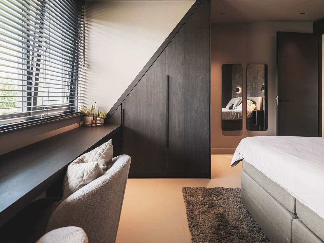 Interieurfoto van een slaapkamer met donkere op maat gemaakte kast onder hellend plafond, raam met jaloezieën en grijze boxspring.